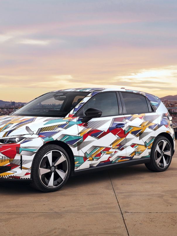 Un VW ID. Polo vopsit în culori vii stă într-o parcare, cu orașul și dealurile în fundal. Vehiculul este acoperit cu un design vibrant, abstract. Cerul va fi parțial înnorat, cu culori de răsărit sau apus.