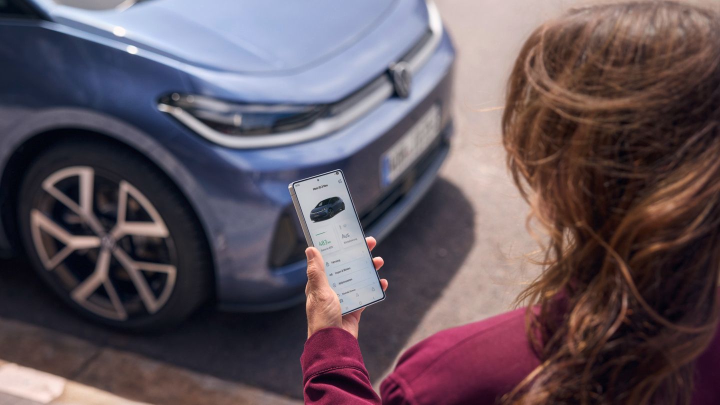 Eine Frau schaut auf ihr Smartphone, vor ihr parkt ein blauer VW ID.3 Neo
