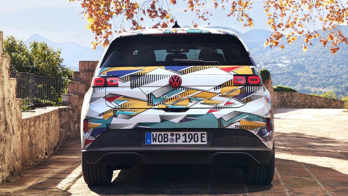 Un ID. Polo stă pe o terasă pavată cu vedere spre un peisaj muntos. Vehiculul are un model geometric colorat și o plăcuță de înmatriculare germană. În fundal, se pot vedea copaci de toamnă și o clădire străveche.