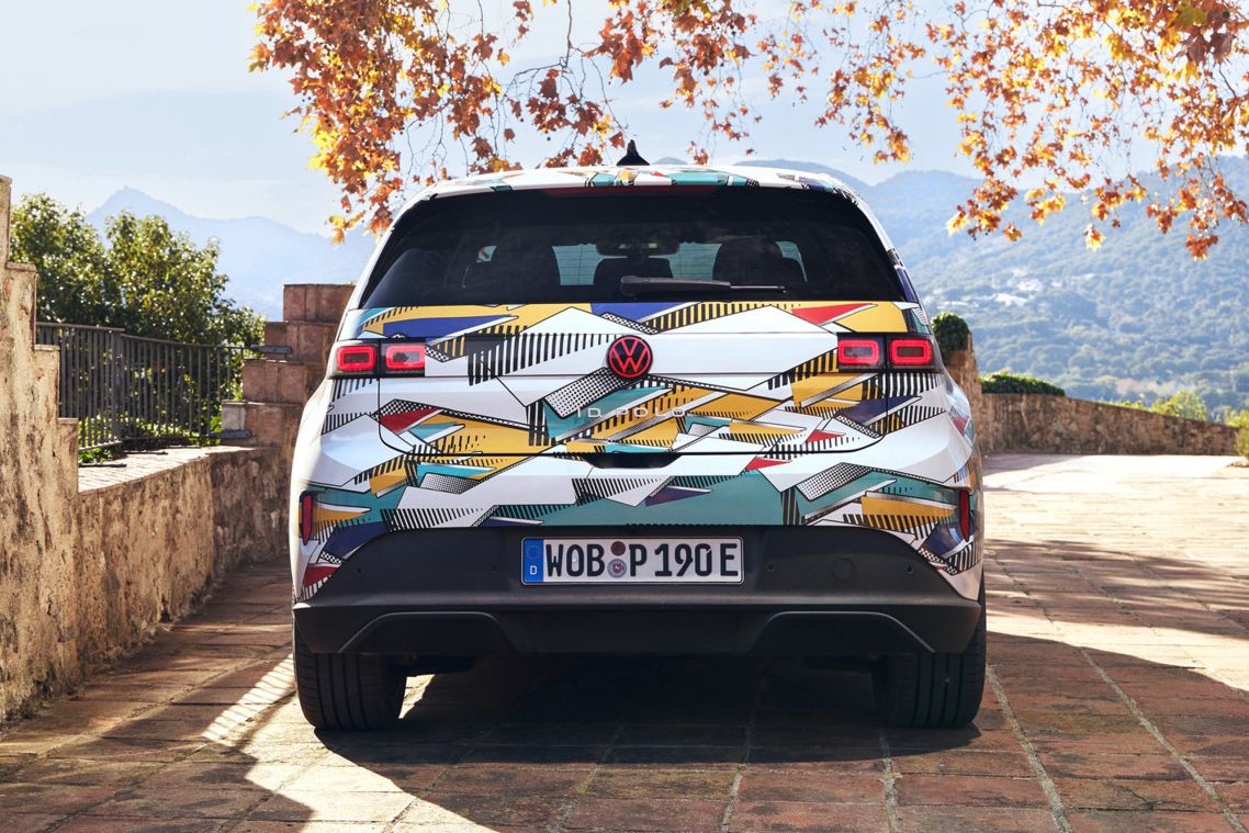 Un ID. Polo stă pe o terasă pavată cu vedere spre un peisaj muntos. Vehiculul are un model geometric colorat și o plăcuță de înmatriculare germană. În fundal, se pot vedea copaci de toamnă și o clădire străveche.