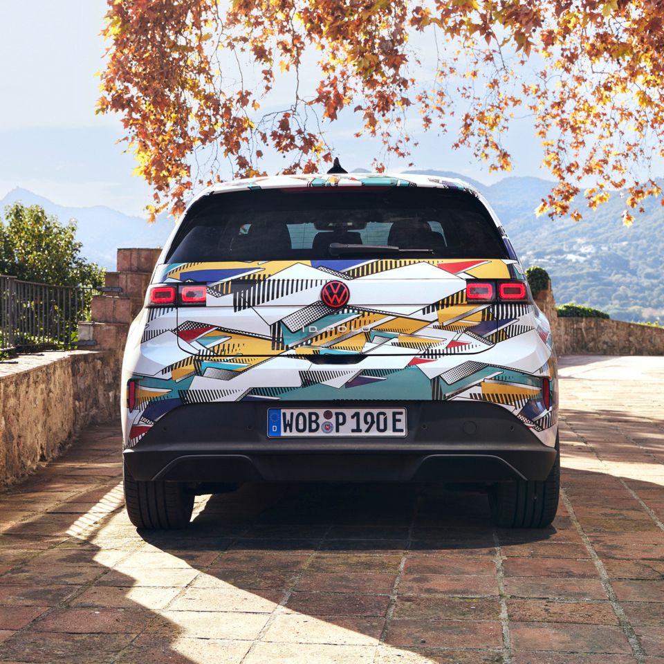 Un ID. Polo stă pe o terasă pavată cu vedere spre un peisaj muntos. Vehiculul are un model geometric colorat și o plăcuță de înmatriculare germană. În fundal, se pot vedea copaci de toamnă și o clădire străveche.