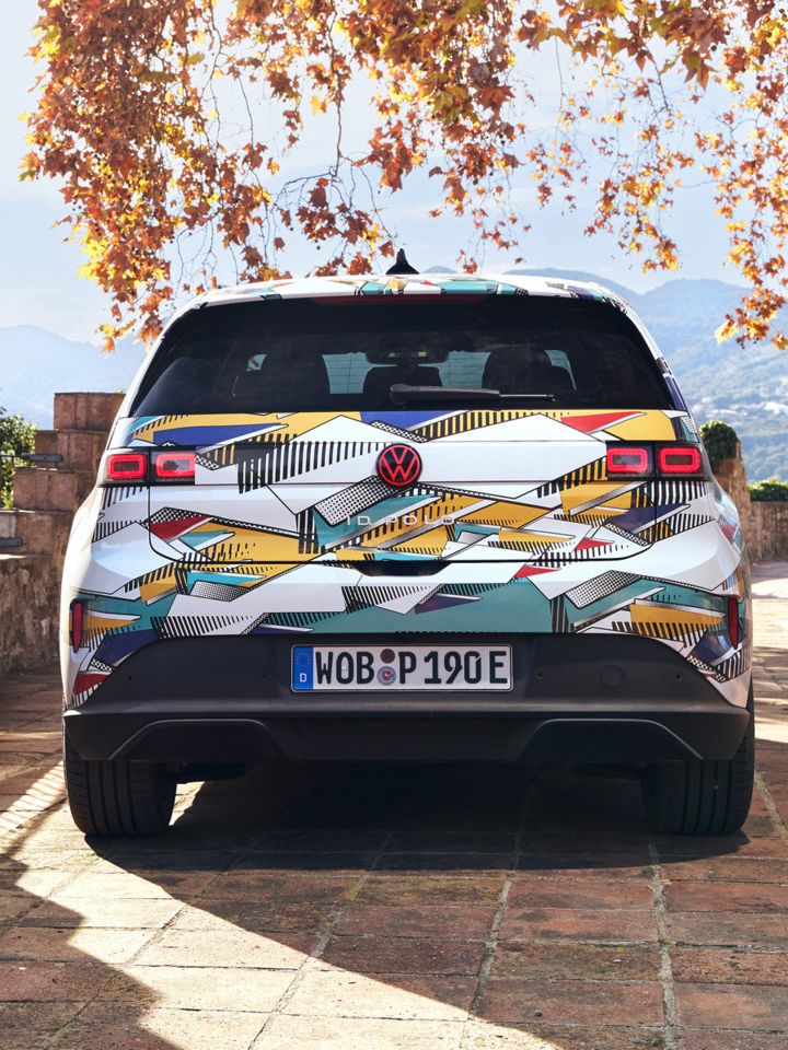 Un ID. Polo stă pe o terasă pavată cu vedere spre un peisaj muntos. Vehiculul are un model geometric colorat și o plăcuță de înmatriculare germană. În fundal, se pot vedea copaci de toamnă și o clădire străveche.