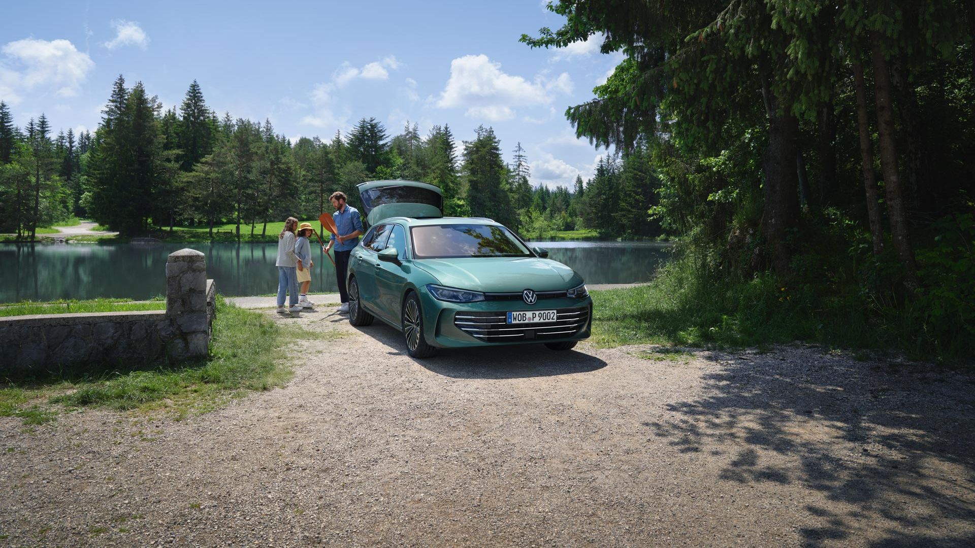 Un VW Passat verde albăstrui parcat lângă un ștrand, cu portbagajul deschis. Un bărbat le împarte jucării celor doi copii.
