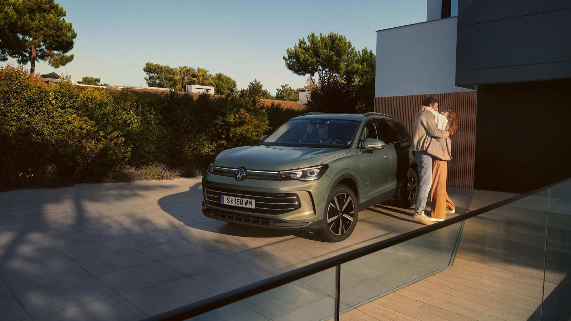 Eine Frau und ein Mann umarmen sich, neben ihnen steht ein VW Tiguan Elegance in der Farbe Cipressino-Grün Metallic