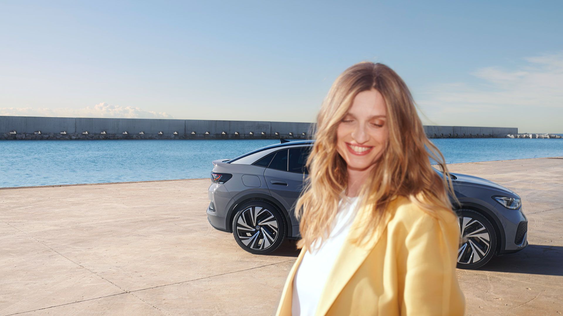O tânără stă zâmbind în fața unui VW ID.5 gri parcat într-un port.