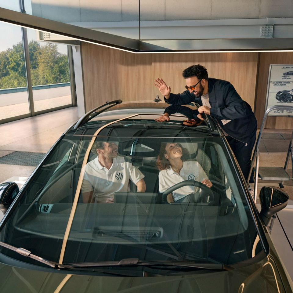 Ein Mann steht auf einer Leiter neben einem VW ID. Cross und winkt durch das Panoramadach den innen sitzenden Personen zu.