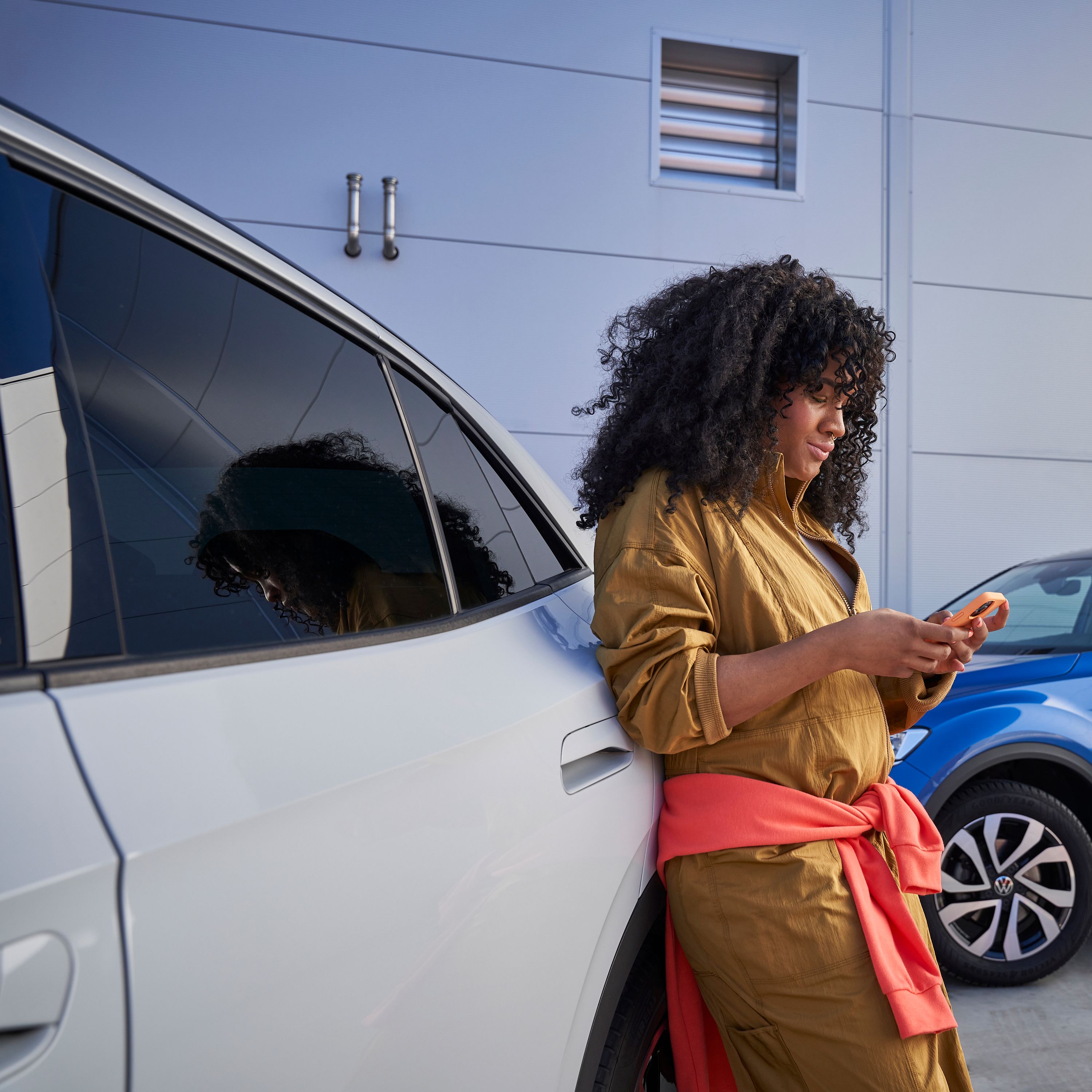 Frau lehnt an Volkswagen und blickt auf ihr Smartphone, im Hintergrund sieht man einen blauen T-Roc