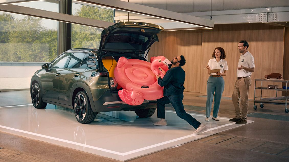 Ein Mann drückt einen aufblasbaren Flamingo in den offenen Kofferraum des VW ID. Cross. Zwei Personen stehen daneben und gucken ihm amüsiert zu.