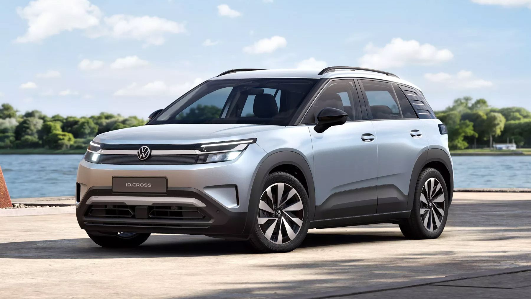 Silberner VW ID. Cross Life mit modernen, kantigen Linien steht auf einer betonierten Fläche an einem Gewässer. Im Hintergrund sind Bäume und ein leicht bewölkter Himmel zu sehen. Das Fahrzeug wirkt vollelektrisch und futuristisch gestaltet.