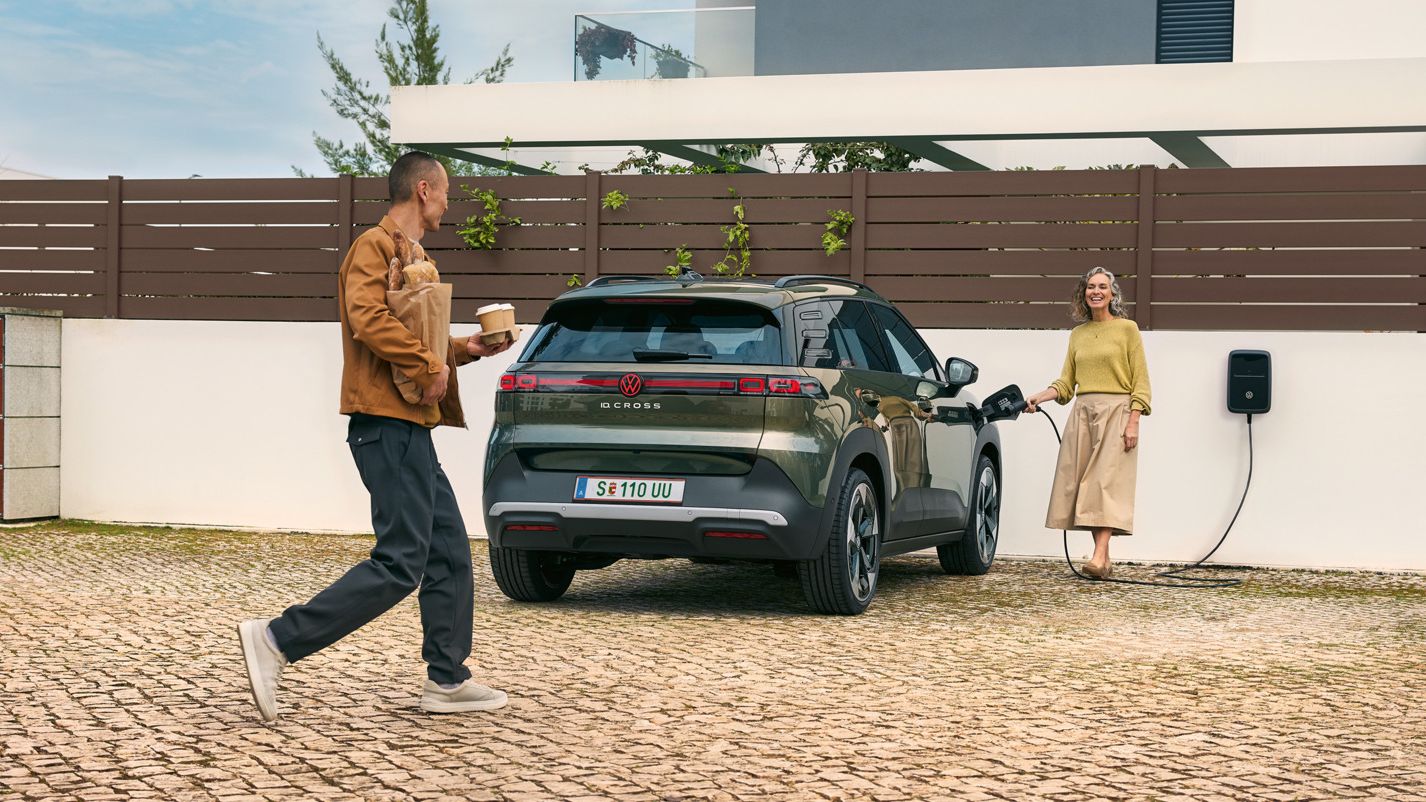 Eine Frau steckt ein Ladekabel einer Wallbox in einen grünen VW ID. Cross, ein Mann geht auf sie zu und hat Kaffeebecher in der Hand und Brot unter dem Arm.