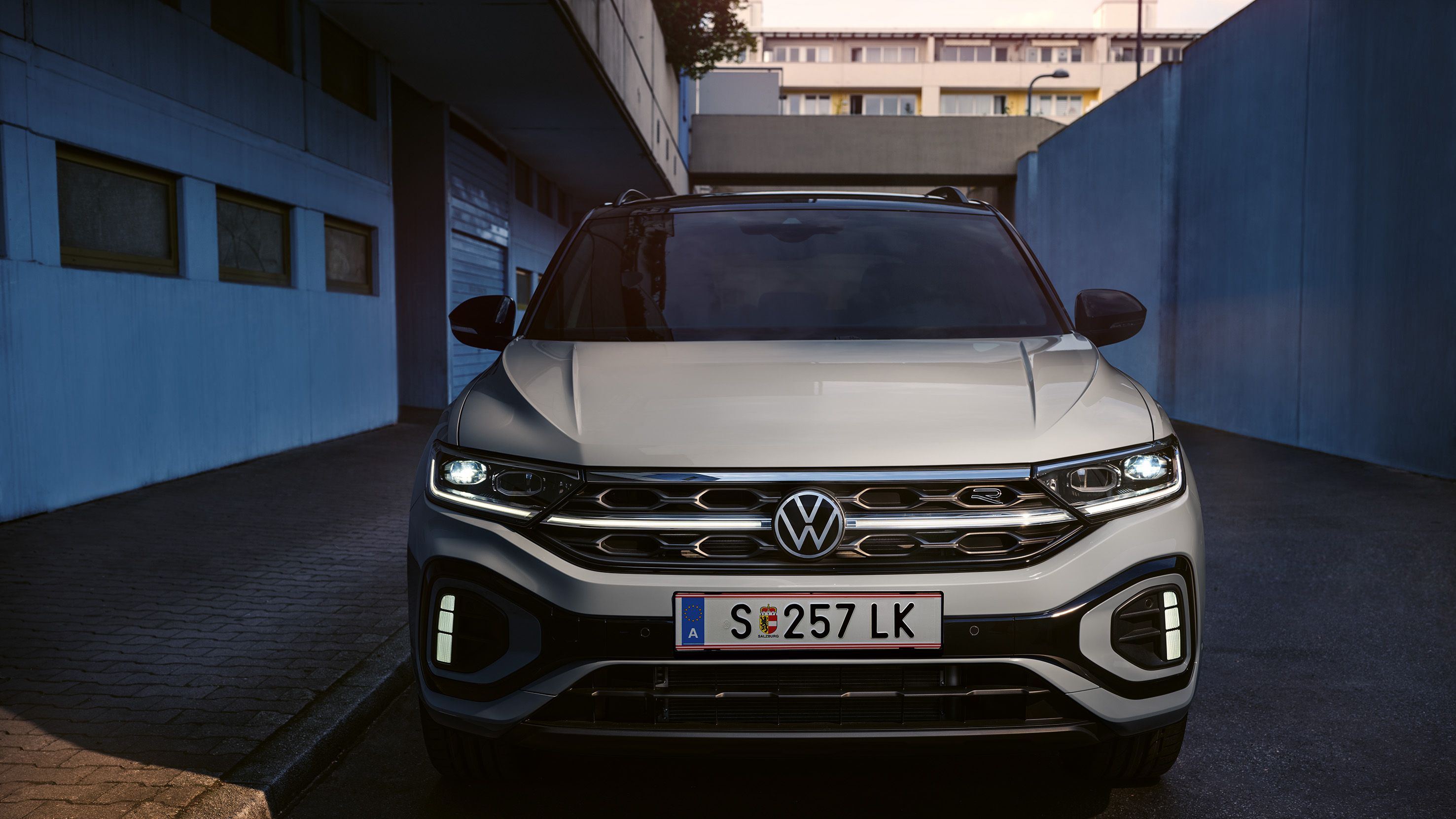 Partea frontală a unui VW T-Roc R-Line de culoare albă, luminile de zi cu leduri și grila radiatorului, iluminată opțional, se aprind