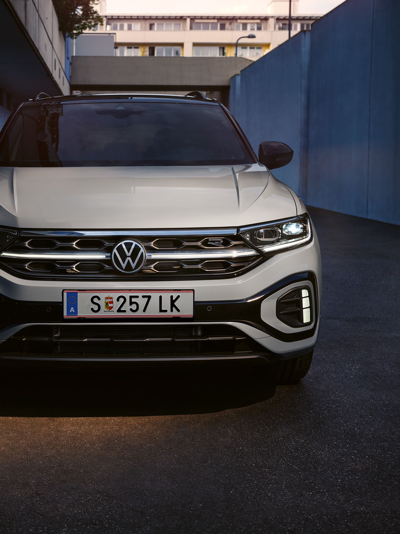 Partea frontală a unui VW T-Roc R-Line de culoare albă, luminile de zi cu leduri și grila radiatorului, iluminată opțional, se aprind