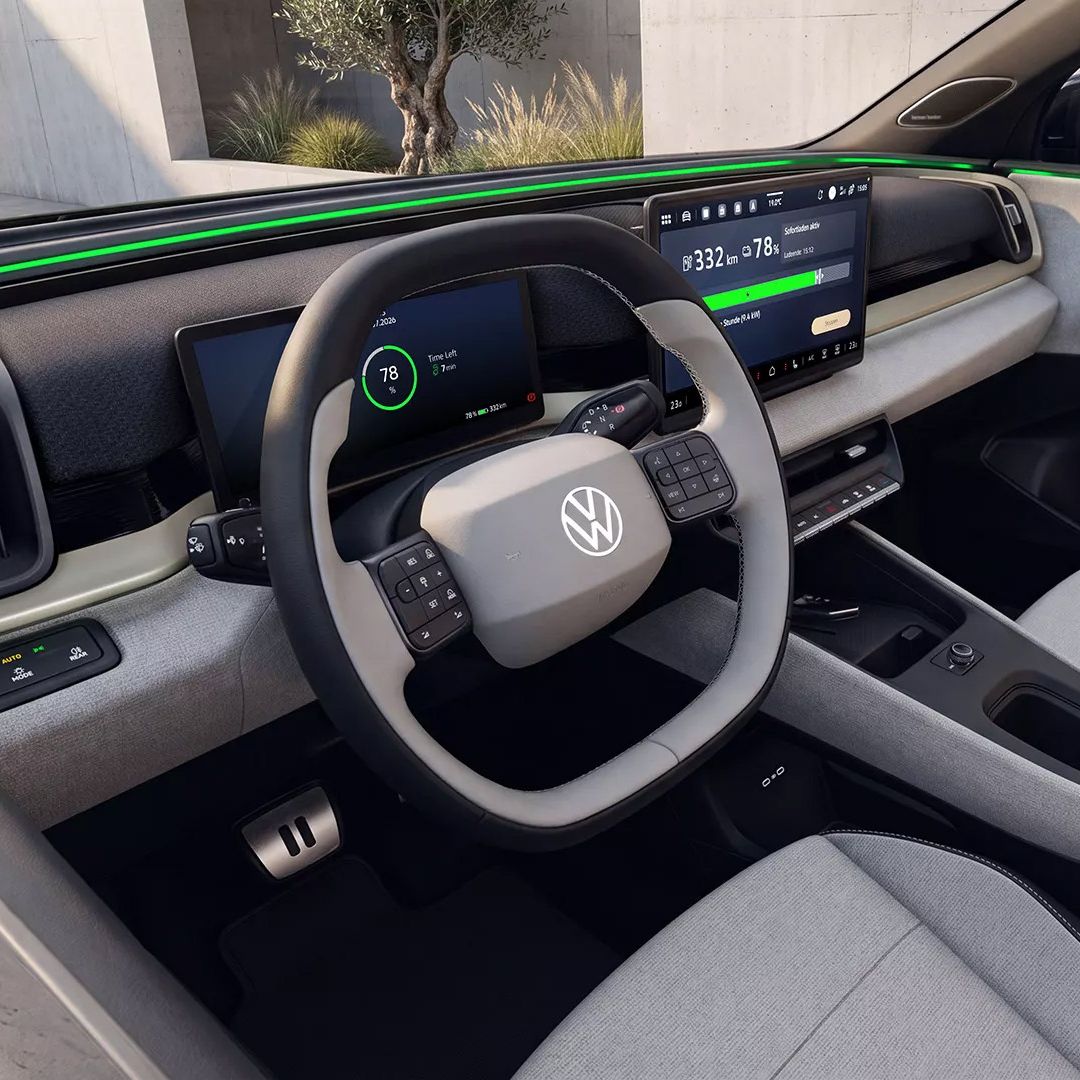 Innenraum eines ID. Cross mit Fokus auf das Cockpit: ein großes zentrales Touchdisplay, ein digitales Kombiinstrument hinter dem Lenkrad und ein reduziertes, hellgraues Armaturenbrett mit wenigen physischen Knöpfen. Das Lenkrad trägt das VW-Logo und ist mit zahlreichen Bedientasten ausgestattet. Im Hintergrund ist eine Ladesäule außerhalb des Fahrzeugs zu sehen.