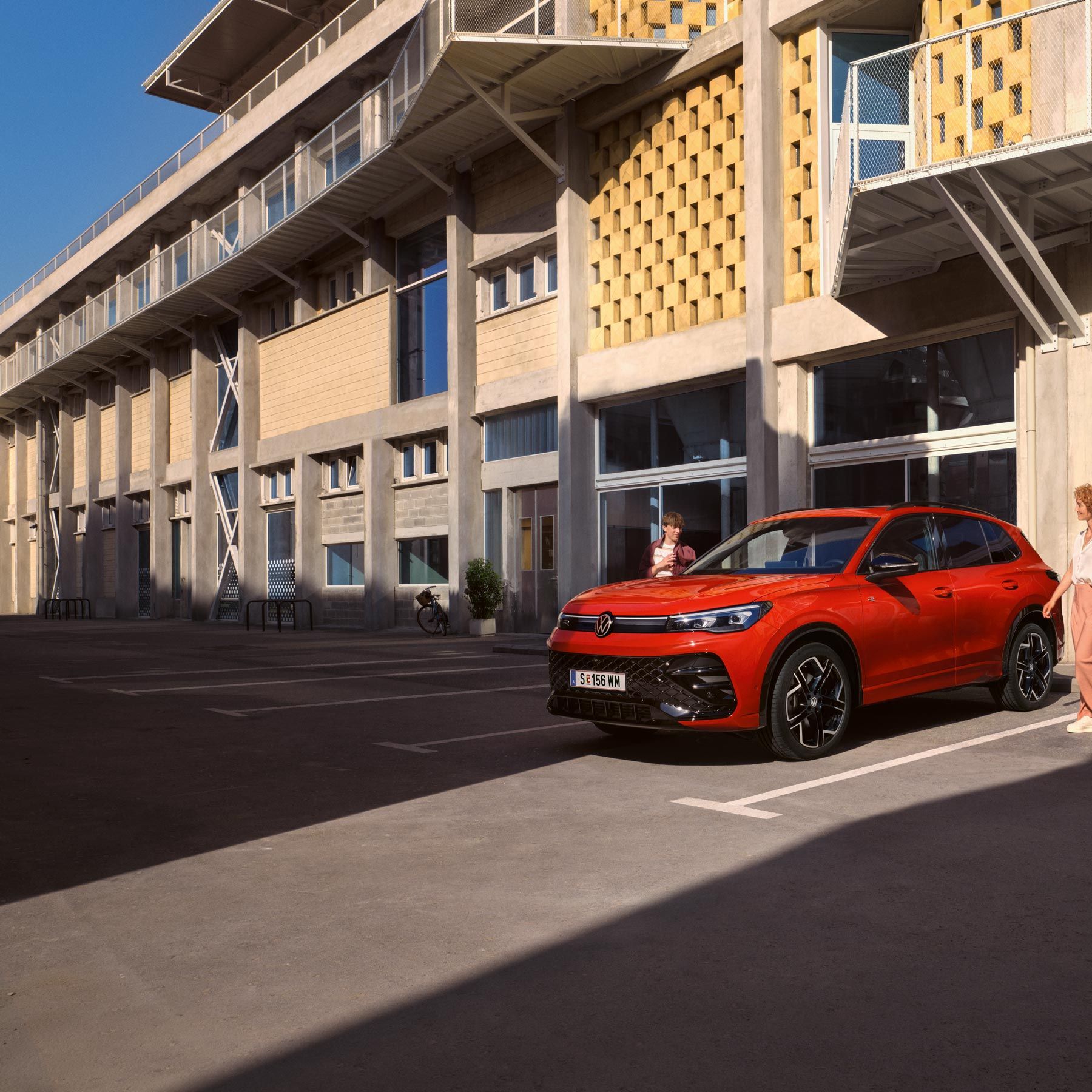 VW Tiguan roșu văzut din lateral în fața unei clădiri, o tânără trece pe lângă partea șoferului.