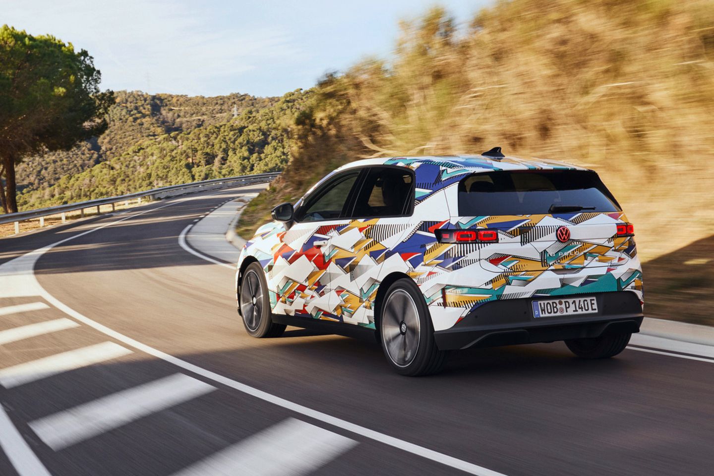 Un ID. Polo cu o vopsea colorată merge pe un drum șerpuit printr-un peisaj deluros. Pe partea dreaptă a drumului poți vedea copaci și tufișuri. Cerul e senin.