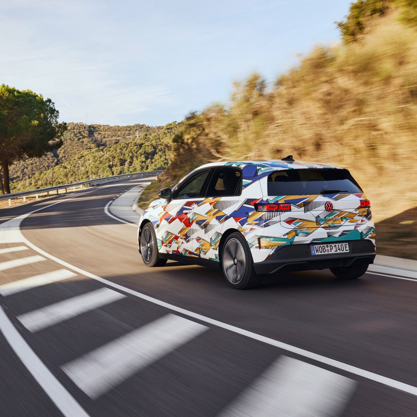 Un ID. Polo cu o vopsea colorată merge pe un drum șerpuit printr-un peisaj deluros. Pe partea dreaptă a drumului poți vedea copaci și tufișuri. Cerul e senin.