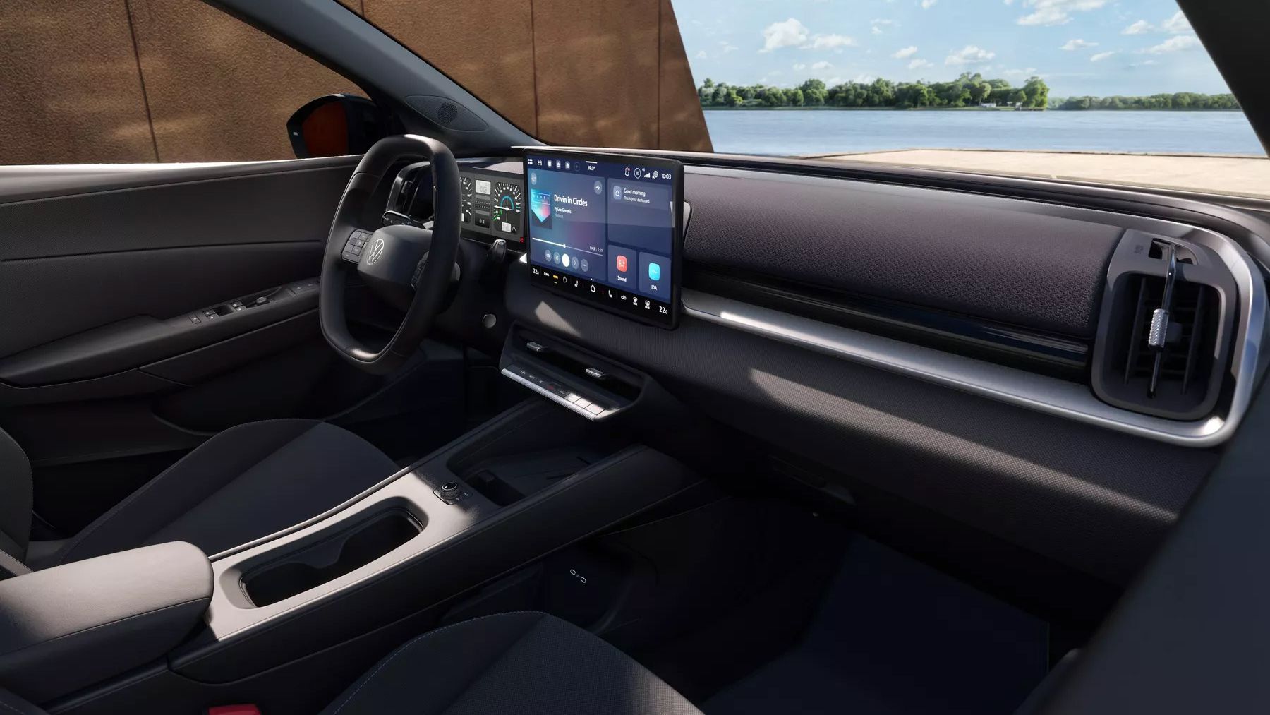 Modernes Cockpit des VW ID. Cross Life mit dunkler Innenausstattung und großem, mittig platziertem Touchscreen für Infotainment. Links ist ein digitales Kombiinstrument hinter dem Lenkrad zu sehen, rechts eine schlichte Armaturentafel mit integrierten Luftdüsen. Durch die Fenster sieht man einen sonnigen Tag mit Wasser und Bäumen im Hintergrund.