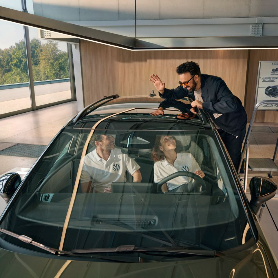 Ein Mann steht auf einer Leiter neben einem VW ID. Cross und winkt durch das Panoramadach den innen sitzenden Personen zu.
