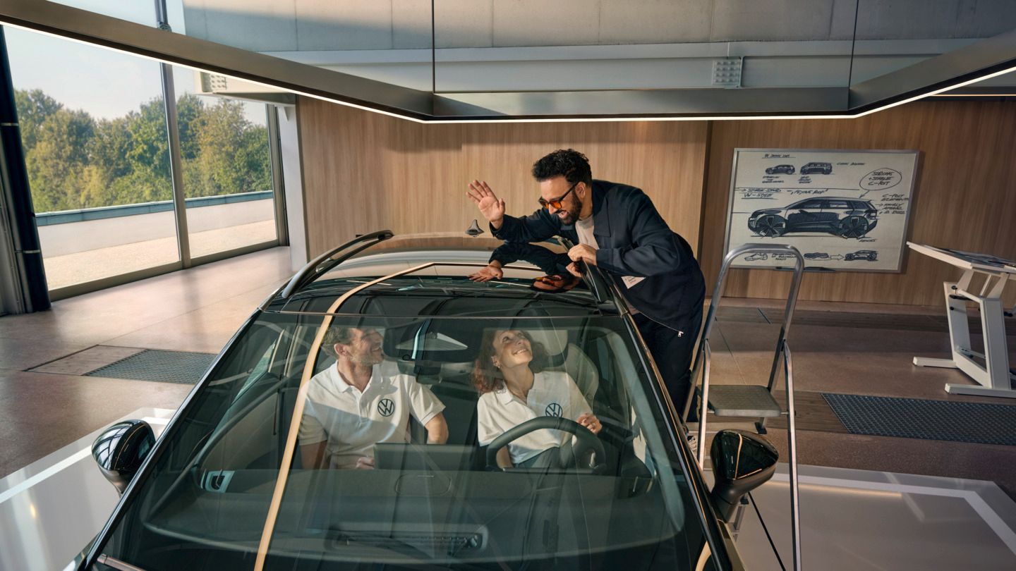Ein Mann steht auf einer Leiter neben einem VW ID. Cross und winkt durch das Panoramadach den innen sitzenden Personen zu.