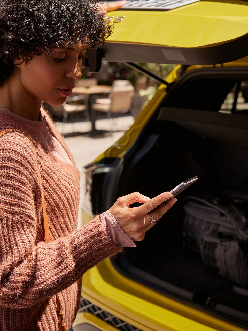 Eine frau schließt den Kofferraum eines gelben VW T-Cross und blickt dabei auf ihr Smartphone. 
