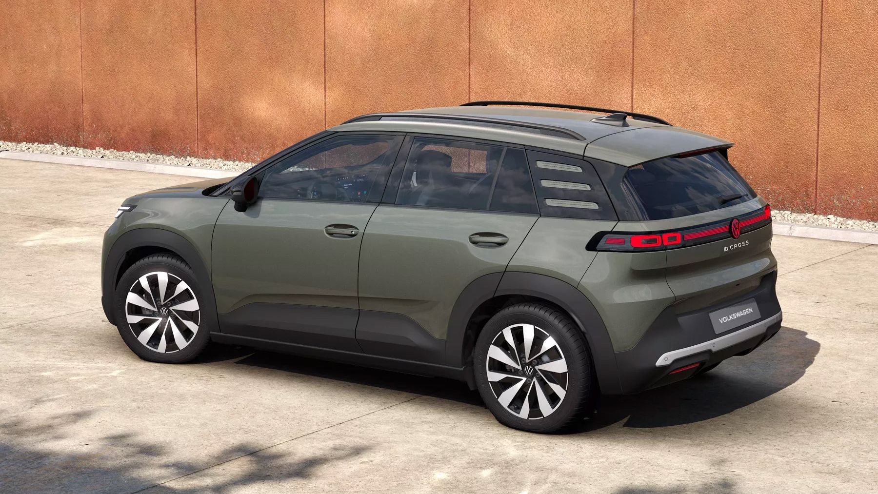 Ein olivgrüner VW ID. Cross Style steht schräg von hinten gesehen auf einem betonierten Platz vor einer orangefarbenen Wand. Das Fahrzeug hat schwarze Dachreling, getönte Scheiben und markante LED-Rückleuchten mit mehreren horizontalen Streifen. Die Felgen sind zweifarbig in Schwarz und Silber gestaltet.
