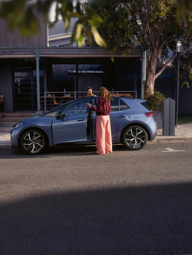 Eine Frau und ein Mann steigen in einen blauen VW ID.3 Neo, der vor einem Café parkt