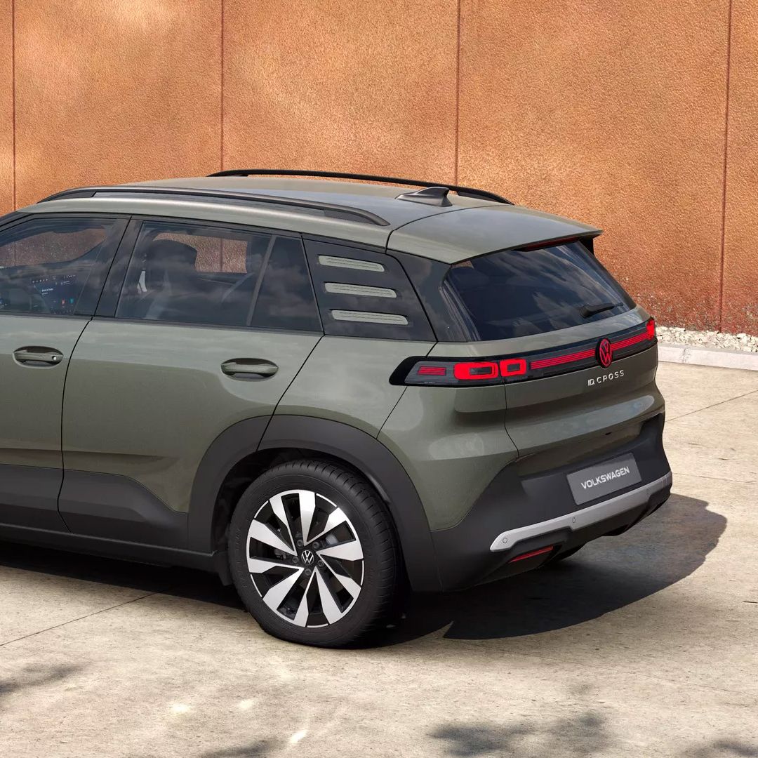 Ein olivgrüner VW ID. Cross Style steht schräg von hinten gesehen auf einem betonierten Platz vor einer orangefarbenen Wand. Das Fahrzeug hat schwarze Dachreling, getönte Scheiben und markante LED-Rückleuchten mit mehreren horizontalen Streifen. Die Felgen sind zweifarbig in Schwarz und Silber gestaltet.