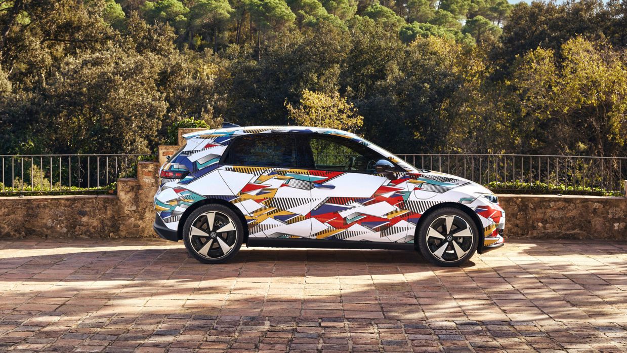 Un ID. Polo cu o imprimare colorată, cu modele geometrice, stă pe un pătrat pietruit. În spatele mașinii se întinde un peisaj împădurit și deluros. Soarele strălucește și vremea este senină.