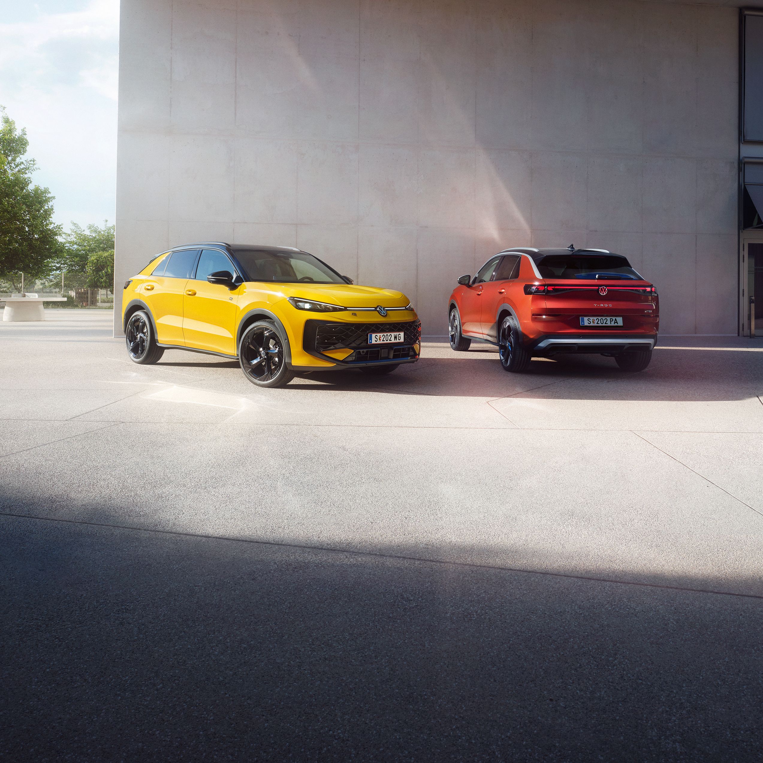 Ein gelber VW T-Roc R-Line und ein roter VW T-Roc Style stehen nebeneinander vor einem modernen Gebäude.