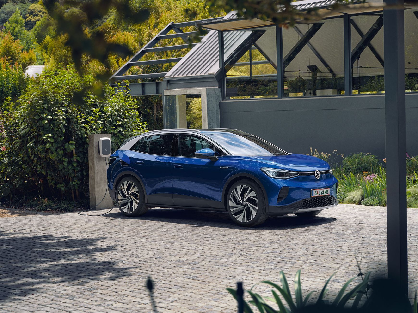 Ein blauer VW ID.4 parkt seitlich vor einem grauen, modernen Haus und lädt an der ID. Charger Wallbox. Im Hintergrund viel grün.
