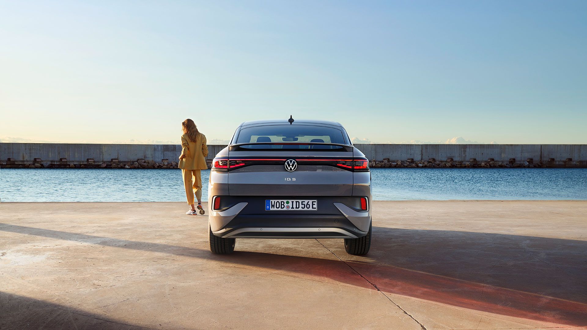 Perspectivă din spate a unui VW ID.5 pe cheiul unui port, o femeie se află lângă mașina electrică