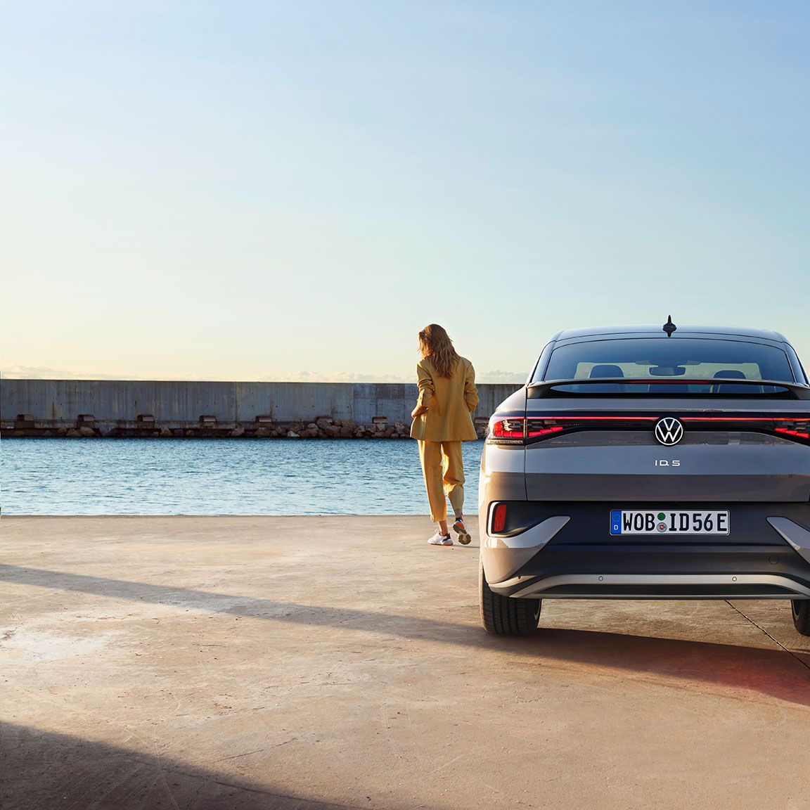 Perspectivă din spate a unui VW ID.5 pe cheiul unui port, o femeie se află lângă mașina electrică
