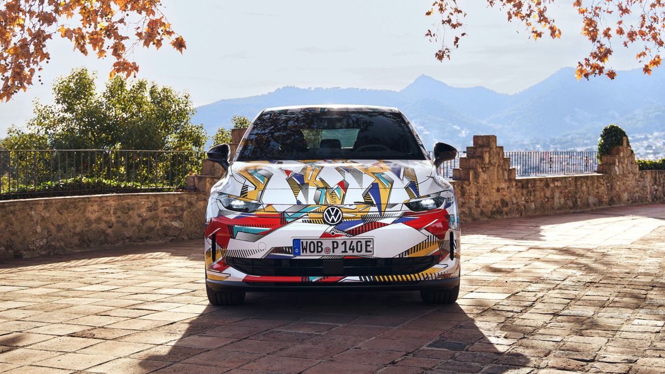 Ein bunt folierter ID. Polo, der auf einem steinernen Weg geparkt ist. Umgeben von herbstlichen Bäumen und mit Bergen im Hintergrund. Die Nummernschilder sind sichtbar.