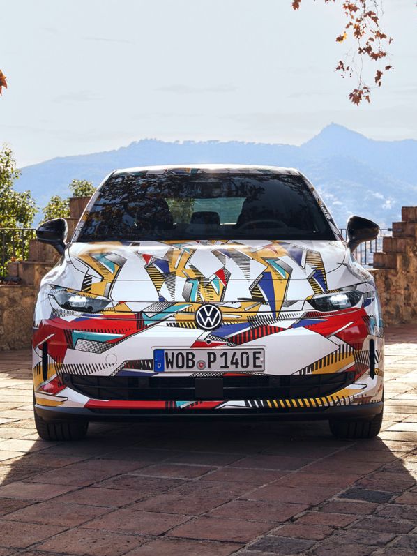 Ein bunt folierter ID. Polo, der auf einem steinernen Weg geparkt ist. Umgeben von herbstlichen Bäumen und mit Bergen im Hintergrund. Die Nummernschilder sind sichtbar.