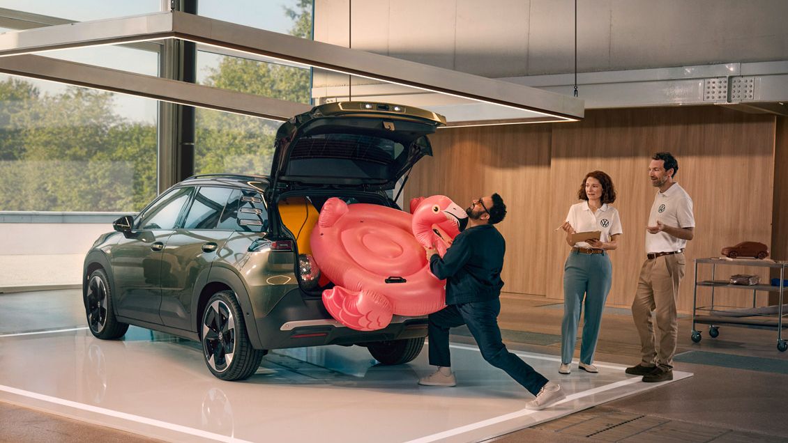 Ein Mann drückt einen aufblasbaren Flamingo in den offenen Kofferraum des VW ID. Cross. Zwei Personen stehen daneben und gucken ihm amüsiert zu.