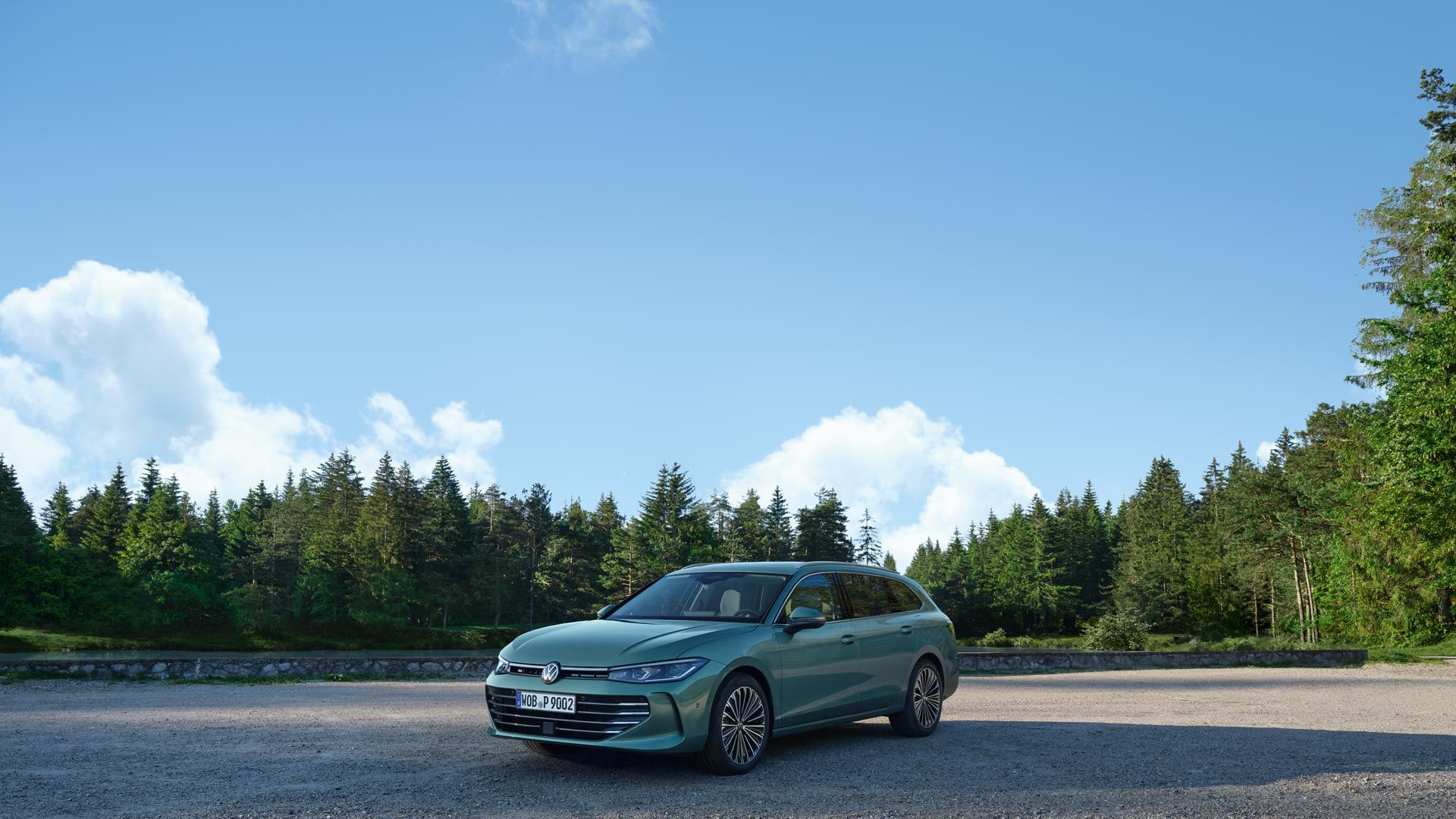 Un VW Passat verde albăstrui pe o suprafață cu pietriș, aflată în vecinătatea unei păduri de conifere.