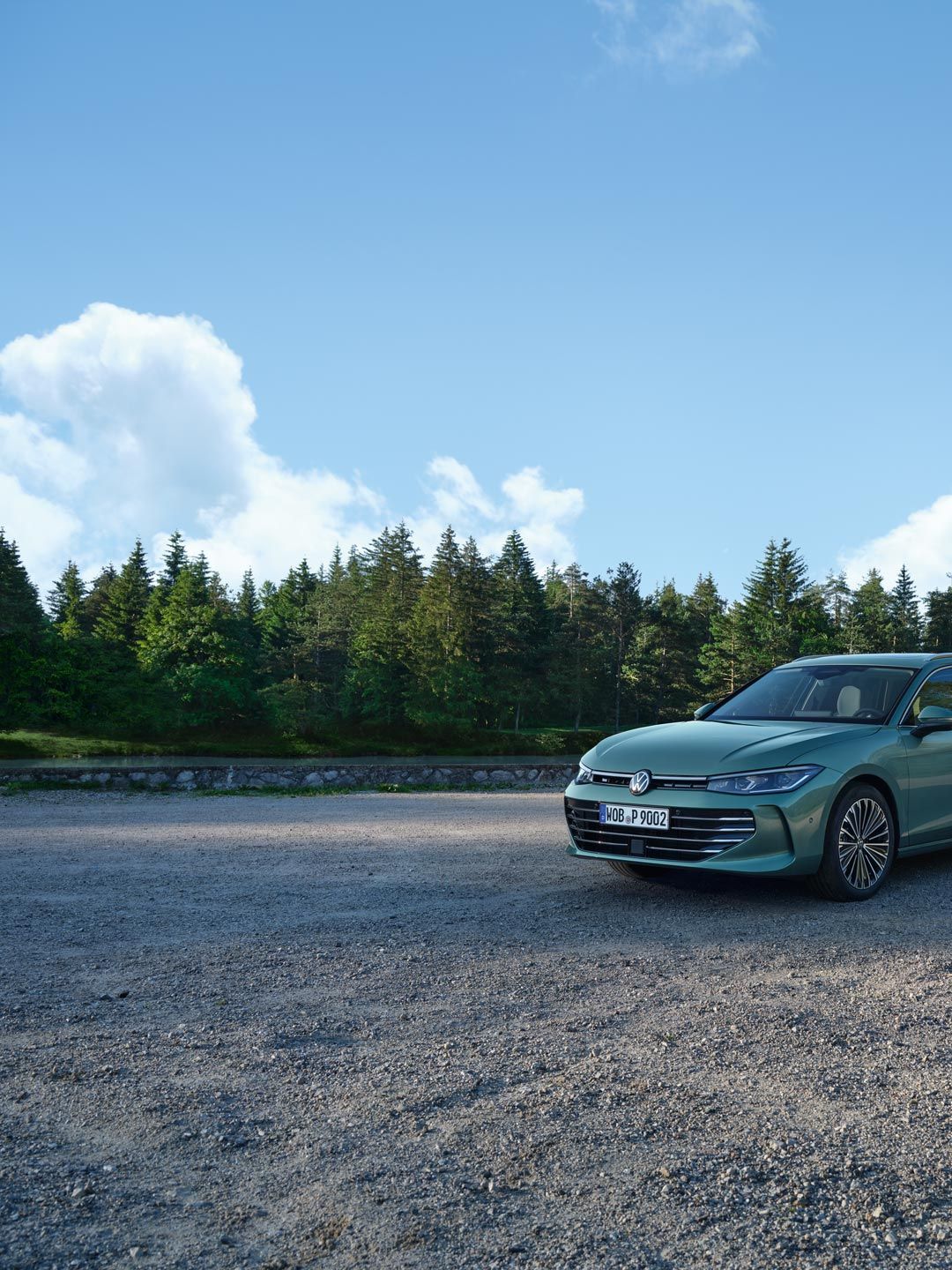Un VW Passat verde albăstrui pe o suprafață cu pietriș, aflată în vecinătatea unei păduri de conifere.