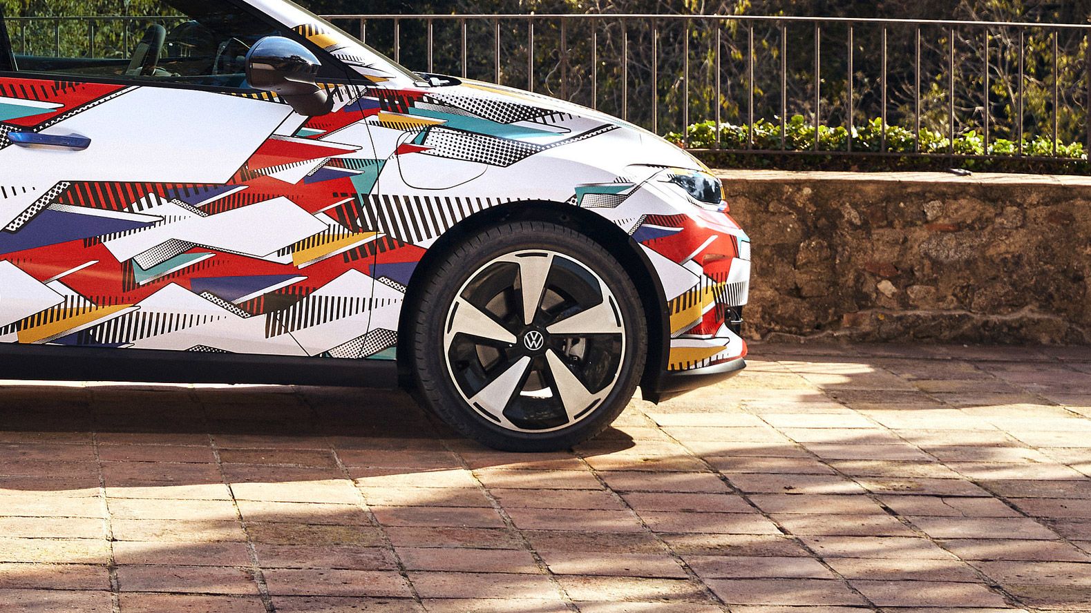 Prim-plan al unui polo ID. cu un design geometric colorat și un logo Volkswagen pe margine. Mașina este parcată pe o alee asfaltată, cu un zid jos de piatră și un peisaj verde în fundal.