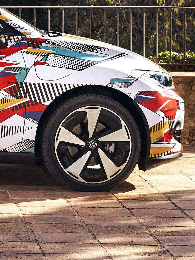 Prim-plan al unui polo ID. cu un design geometric colorat și un logo Volkswagen pe margine. Mașina este parcată pe o alee asfaltată, cu un zid jos de piatră și un peisaj verde în fundal.