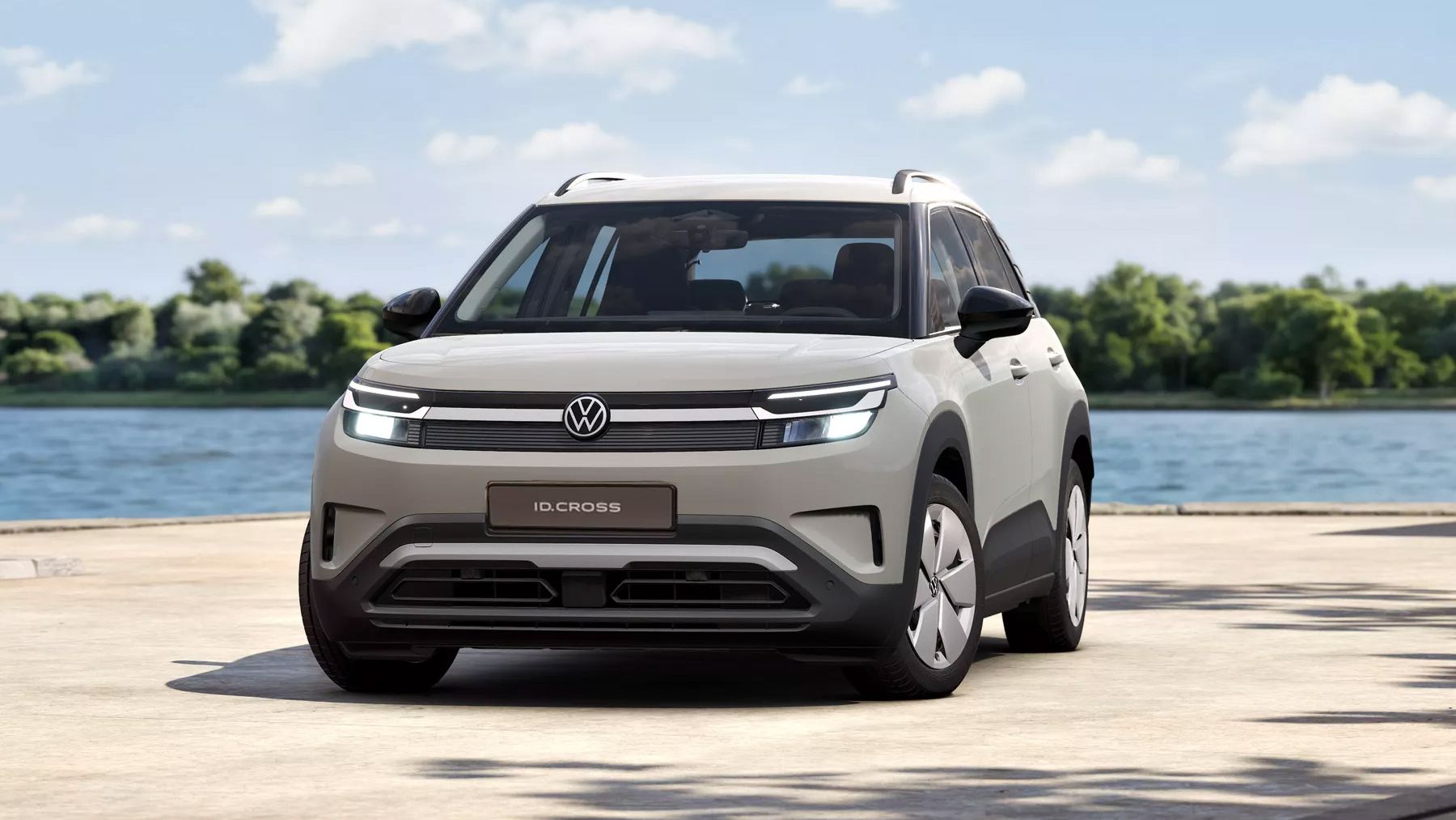 Ein heller, moderner VW ID. Cross steht auf einer Uferpromenade vor einem See, im Hintergrund Bäume und blauer Himmel mit wenigen Wolken. Das Auto ist frontal leicht schräg zu sehen, mit LED-Frontscheinwerfern und VW-Logo in der Mitte.